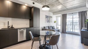 A modern kitchen with dark cabinets, a dishwasher, and a round dining table with chairs. There is a sofa and a window with curtains in the living area.