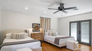 Une chambre avec deux lits, un ventilateur de plafond, une table de chevet, un tableau encadré et une fenêtre avec des stores.