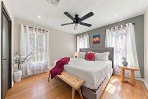 A bedroom with a bed, a bench, a bedside table, a ceiling fan, and a window with curtains.