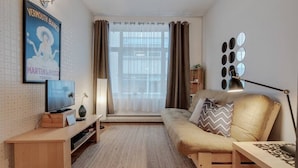 A cozy living room with a sofa, a TV on a wooden stand, a small table, and a lamp. There is a window with curtains, a poster on the wall, and a mirror on the wall.