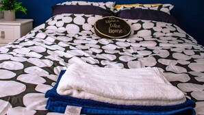 Un lit bien fait, recouvert d’une couvre-lit à motifs floraux noir et blanc, un oreiller orné du message « Il n’y a pas de meilleur endroit que chez soi », et une serviette blanche posée sur le lit.