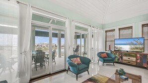 A spacious living area with a large sliding glass door leading to an outdoor deck, a television on a wooden stand, and two teal armchairs. - 3BR Lakefront Retreat w/ Modern Decor + Fire Pit | It Doesn't Get Any Better (Destin)