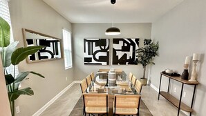 A dining area with a glass table surrounded by wicker chairs, a large abstract painting on the wall, and a potted plant. - Luxury Family Estate | Orlando Pool and Game Room (Kissimmee)