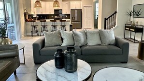 A modern living room with a grey sofa, two round marble tables, and a kitchen in the background. - Luxury Family Estate | Orlando Pool and Game Room (Kissimmee)