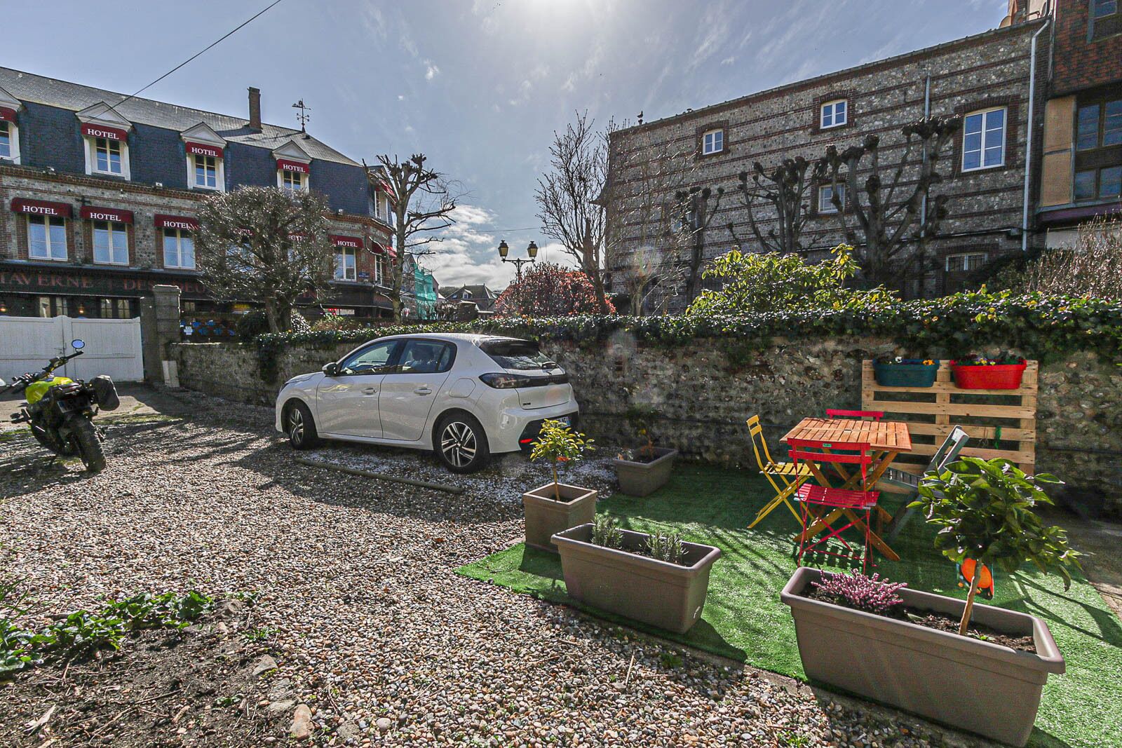 A white car parked in a gravel driveway with a garden area featuring potted plants and outdoor furniture.