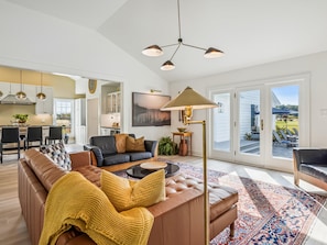 A spacious living room with a sofa, coffee table, and a patterned rug. There is a dining area with chairs and a table, and a kitchen in the background.