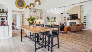 A spacious living area with a wooden dining table, black chairs, and a brown sofa. There is a kitchen area with white cabinets and a large window. - Minutes from Downtown St. Michaels | Pool | Hot Tub | Pickle Ball Court (Saint Michaels)
