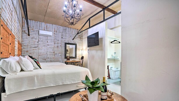 Een slaapkamer met een bed, een houten hoofdbord, een kroonluchter, een spiegel, een tafeltje met een plant en fles, en een wasmand op de achtergrond.