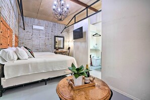 Ein Schlafzimmer mit einem Bett, einem hölzernen Kopfteil, einer Chandelierleuchte, einem Spiegel, einem kleinen Tisch mit Pflanze und Flasche sowie einem Wäschekorb im Hintergrund.
