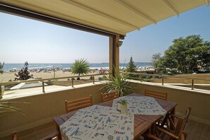 Un área de comedor al aire libre con una mesa para dos, con vista a una playa con sombrillas y gente.