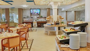 Een restaurantruimte in een hotel, met meerdere tafels, stoelen, een buffet en een televisiescherm.