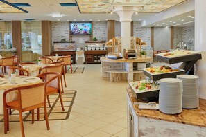 A hotel dining area with multiple tables, chairs, a buffet station, and a television screen.