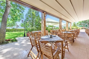 An outdoor dining area with wooden tables and chairs, a covered walkway, and a view of the sea.