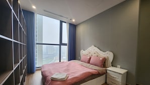 Un dormitorio con un ventanal grande, una cama con ropa de cama rosa, una mesita de noche blanca y un armario de madera.