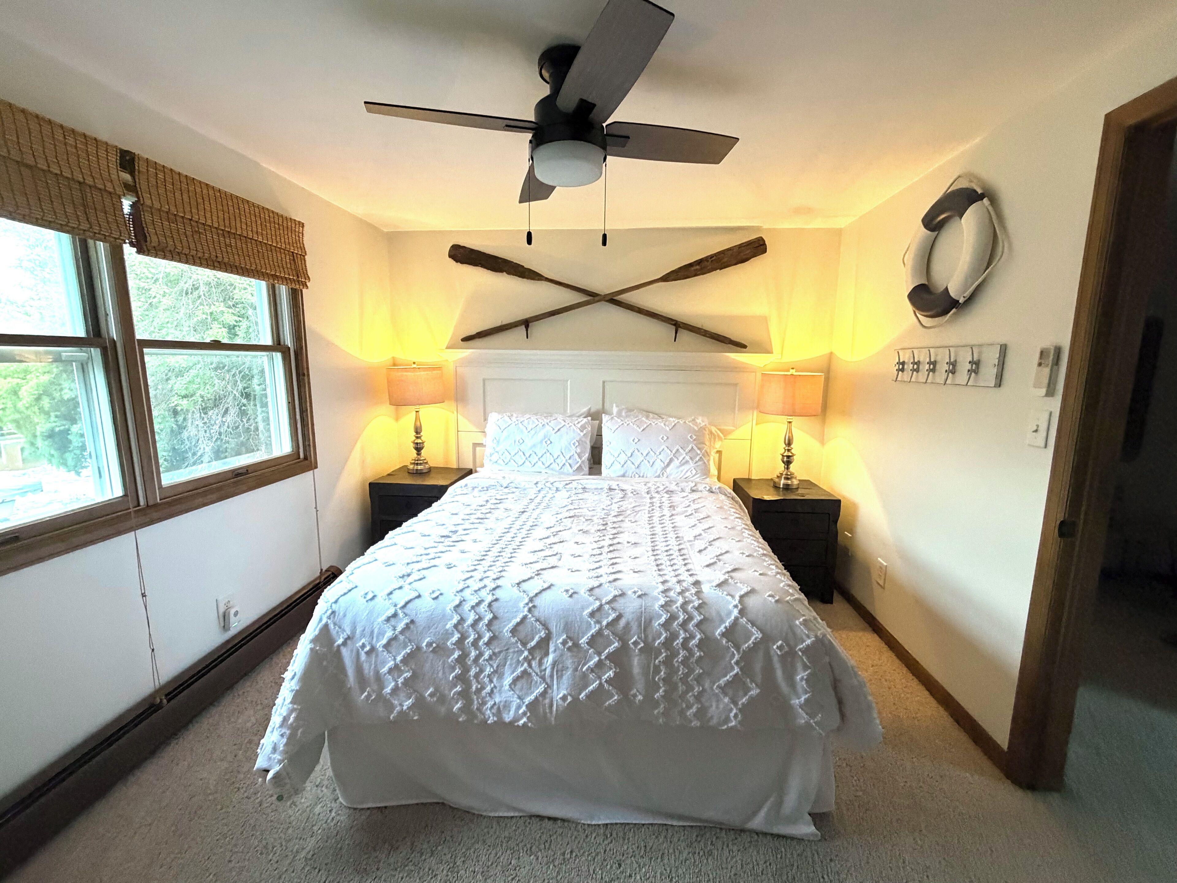 A bedroom with a bed, two bedside tables with lamps, a ceiling fan, a window with blinds, and a wall-mounted coat rack.