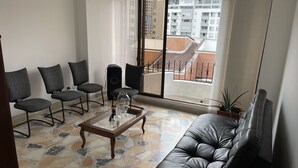 Ett vardagsrum med en svart lädersoffa, tre grå fåtöljer, ett glasbord och ett stort fönster med utsikt över byggnader och en balkong.