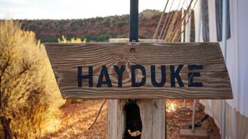 Uma placa de madeira com o nome 'HAYDUKE' fixada num poste, com um painel solar acima, com um edifício e paisagem desértica ao fundo.
