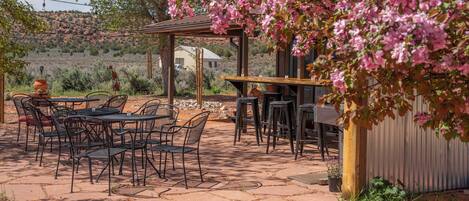 Une terrasse extérieure avec des tables et des chaises, une pergola ornée de fleurs roses et une vue sur un paysage désertique.
