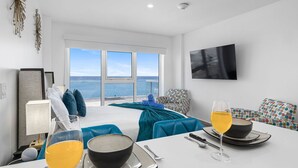 Una habitación de hotel moderna con una cama grande, una mesa de comedor con alimentos para el desayuno, un televisor y vista al mar.