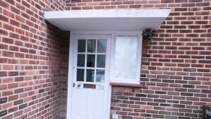 A brick building with a white door marked '20', a small window, and a visible garden hose. - South Harrow Apartment With Free Parking (Harrow)