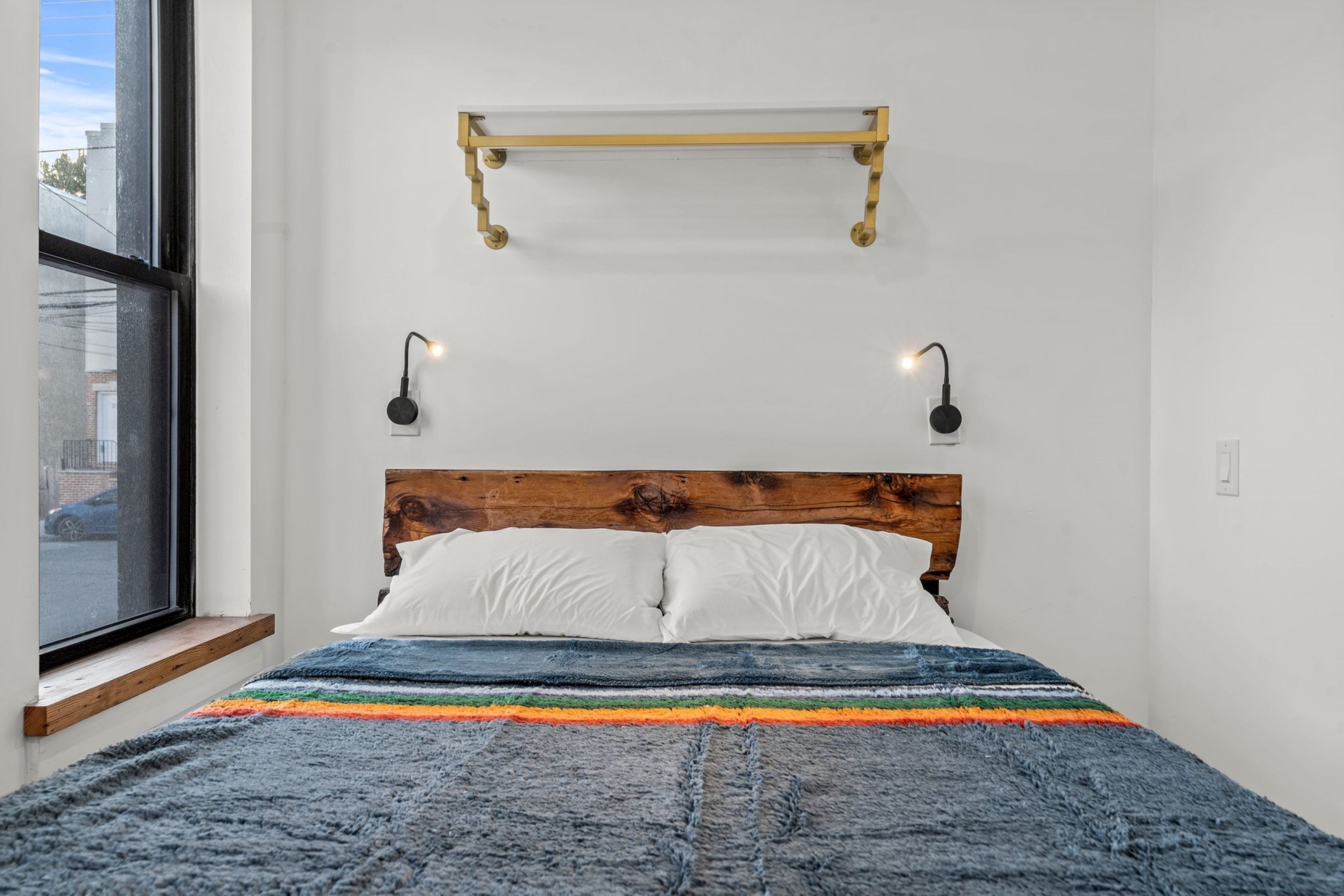 A neatly made bed with a wooden headboard, white pillows, and a colorful striped blanket. There is a small shelf above the bed and two wall-mounted lamps on either side.