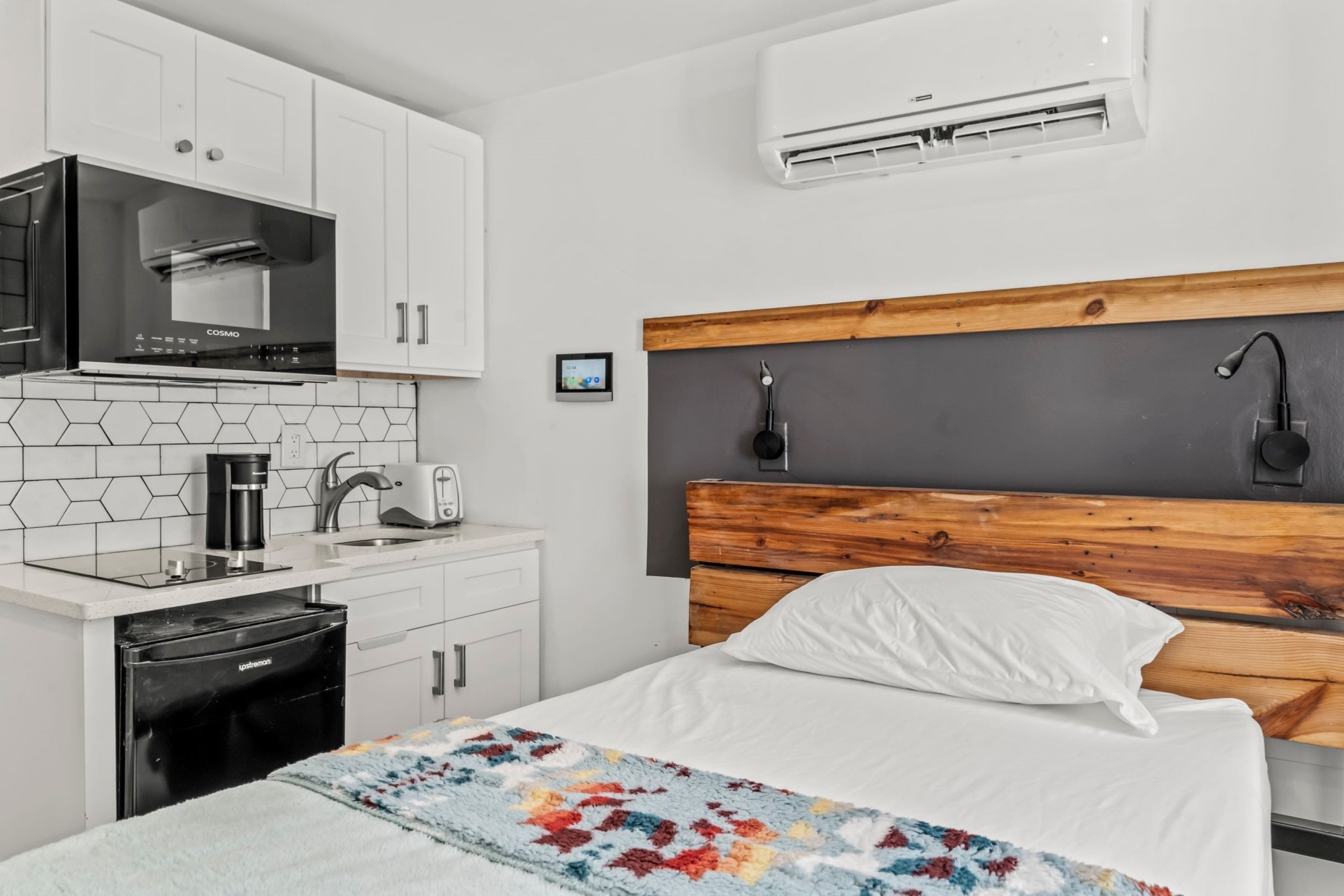 A modern hotel room with a kitchen area, a bed with a wooden headboard, and an air conditioning unit.