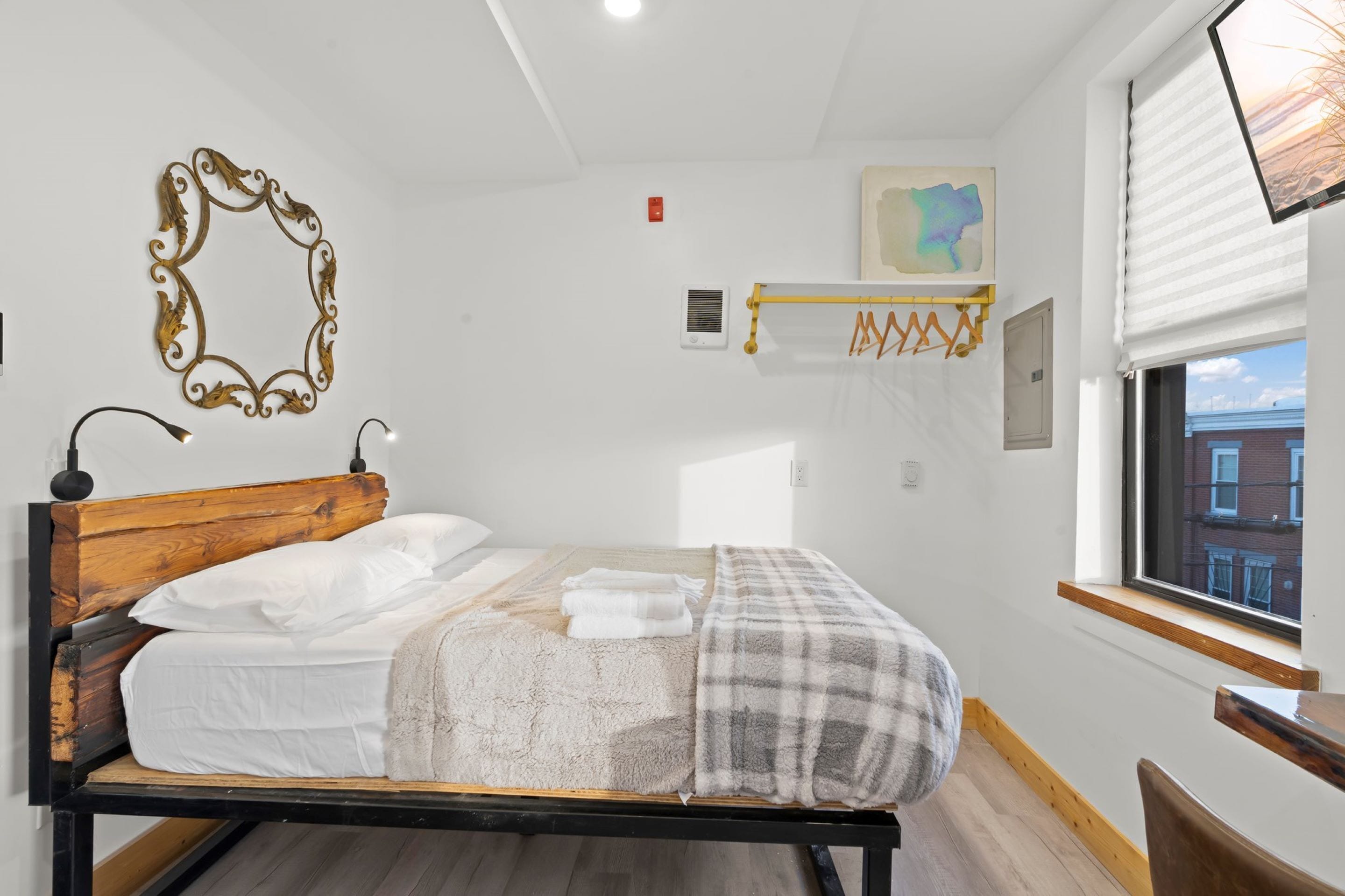 A neatly made bed with white linens, a wooden headboard, and a plaid blanket. There is a wall-mounted mirror with an ornate frame, a small shelf with a painting, and a window with a view of buildings.