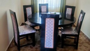 A dining area with a round table surrounded by six chairs, each with a patterned cushion. The room has a tiled floor and a window with green curtains.