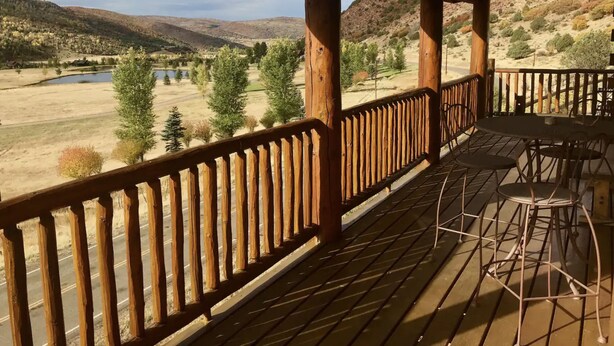A wooden deck with metal chairs and tables, overlooking a valley with trees and a body of water.