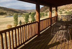 Una terraza de madera con sillas y mesas de metal, con vista a un valle con árboles y un cuerpo de agua.