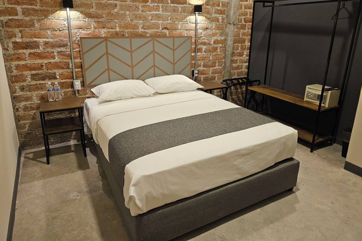 Quarto moderno com parede de tijolos, cama com cabeceira cinza, mesas de cabeceira com abajures e uma pequena mesa de madeira com um rádio.