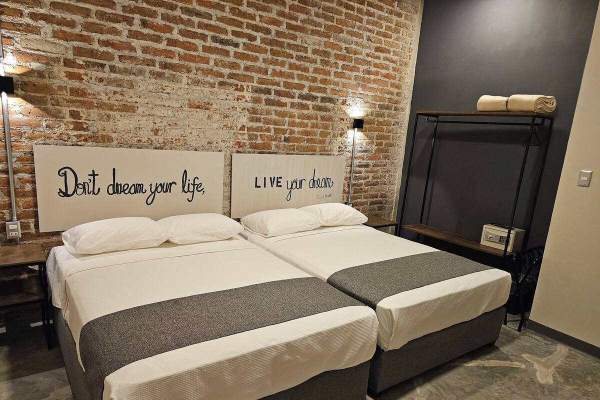 Uma cama de casal com colcha cinza e branca, duas mesas de cabeceira, uma parede de tijolos e pôsteres motivacionais nas paredes.