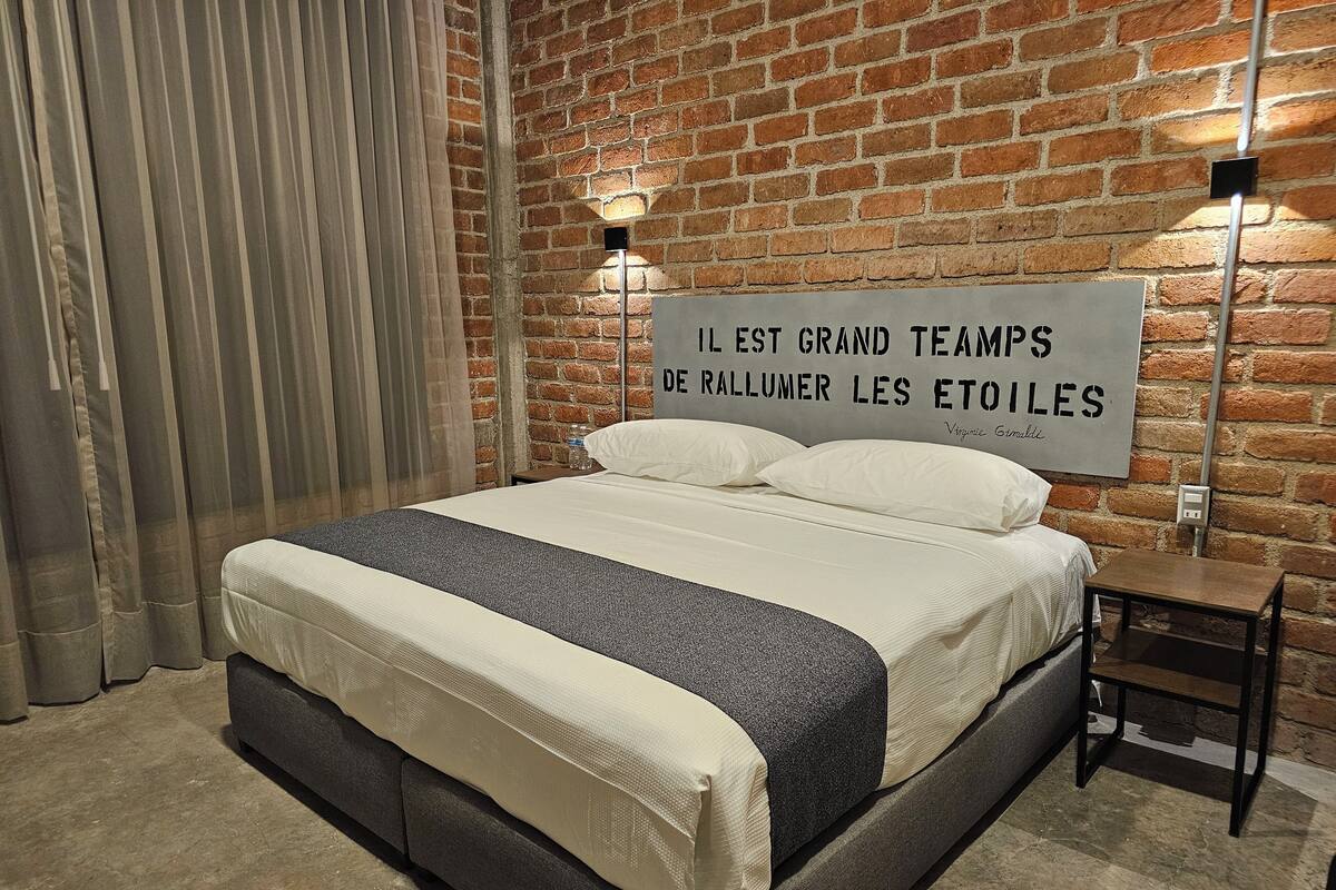 Quarto com parede de tijolos, cama com roupa de cama branca e cinza, mesa de cabeceira pequena e um letreiro com texto em francês.