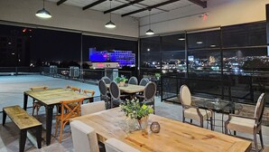 An outdoor dining area with tables and chairs, large windows offering city views, and a high ceiling with exposed beams. - HOTEL THE ROOF (León)