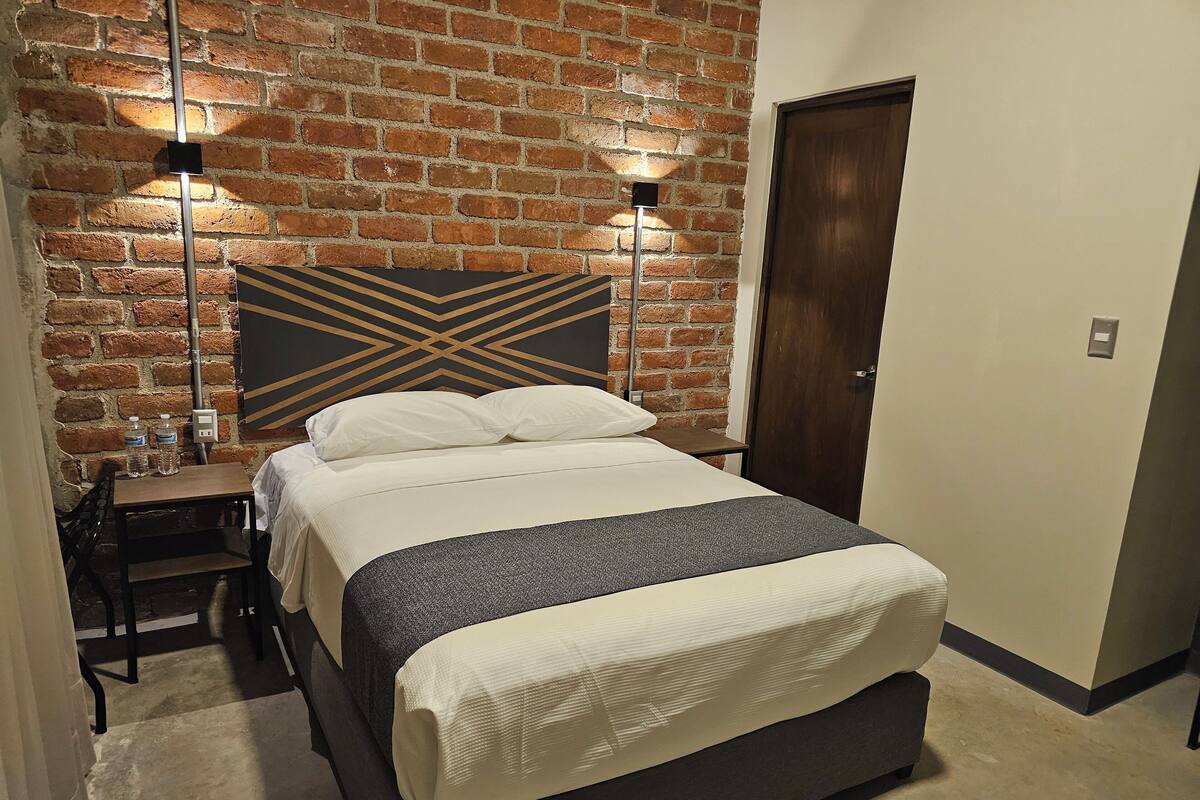Quarto de hotel com parede de tijolos, uma cama com roupa de cama branca, duas mesas de cabeceira com abajures e uma porta de madeira.