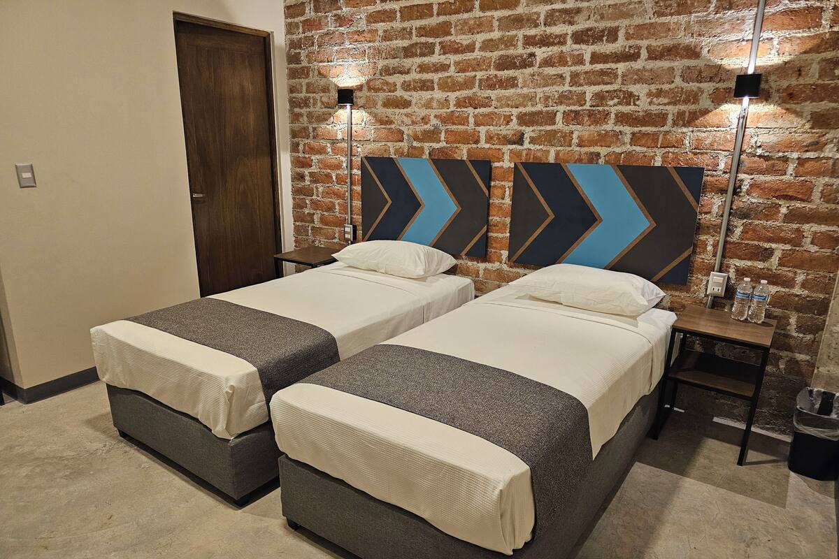 Quarto de hotel com duas camas, uma parede de tijolos e uma mesinha de cabeceira com duas garrafas de água.