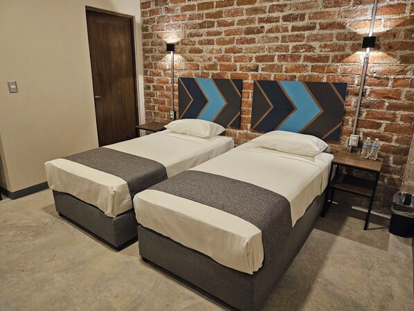 Quarto de hotel com duas camas, uma parede de tijolos e uma mesinha de cabeceira com duas garrafas de água.