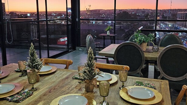 A restaurant with a city view at sunset, featuring a wooden table set for dining, chairs, and a small Christmas tree. - HOTEL THE ROOF (León)