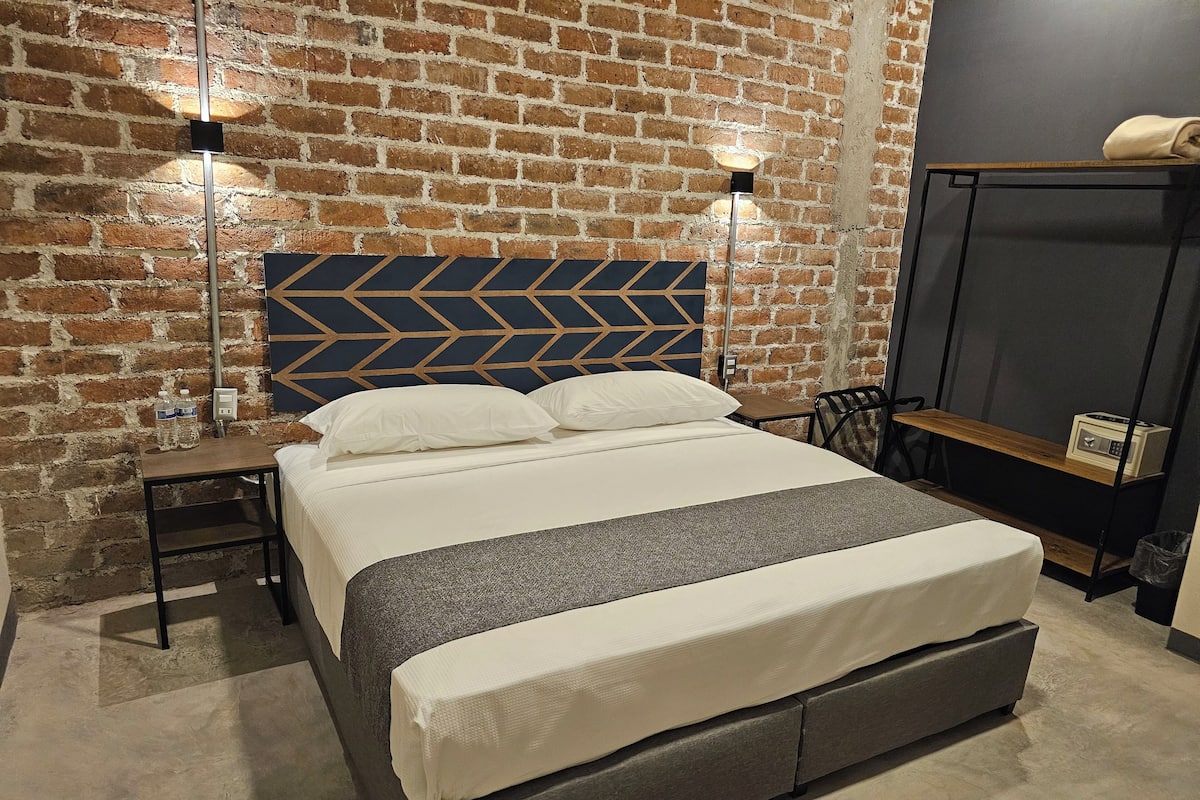 Quarto de hotel moderno com parede de tijolos, cama com cabeceira geométrica, mesas de cabeceira e uma pequena escrivaninha com um rádio.