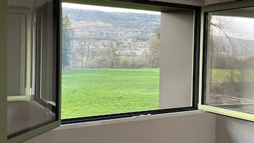 A room with a large window offering a view of a green field and a distant town.