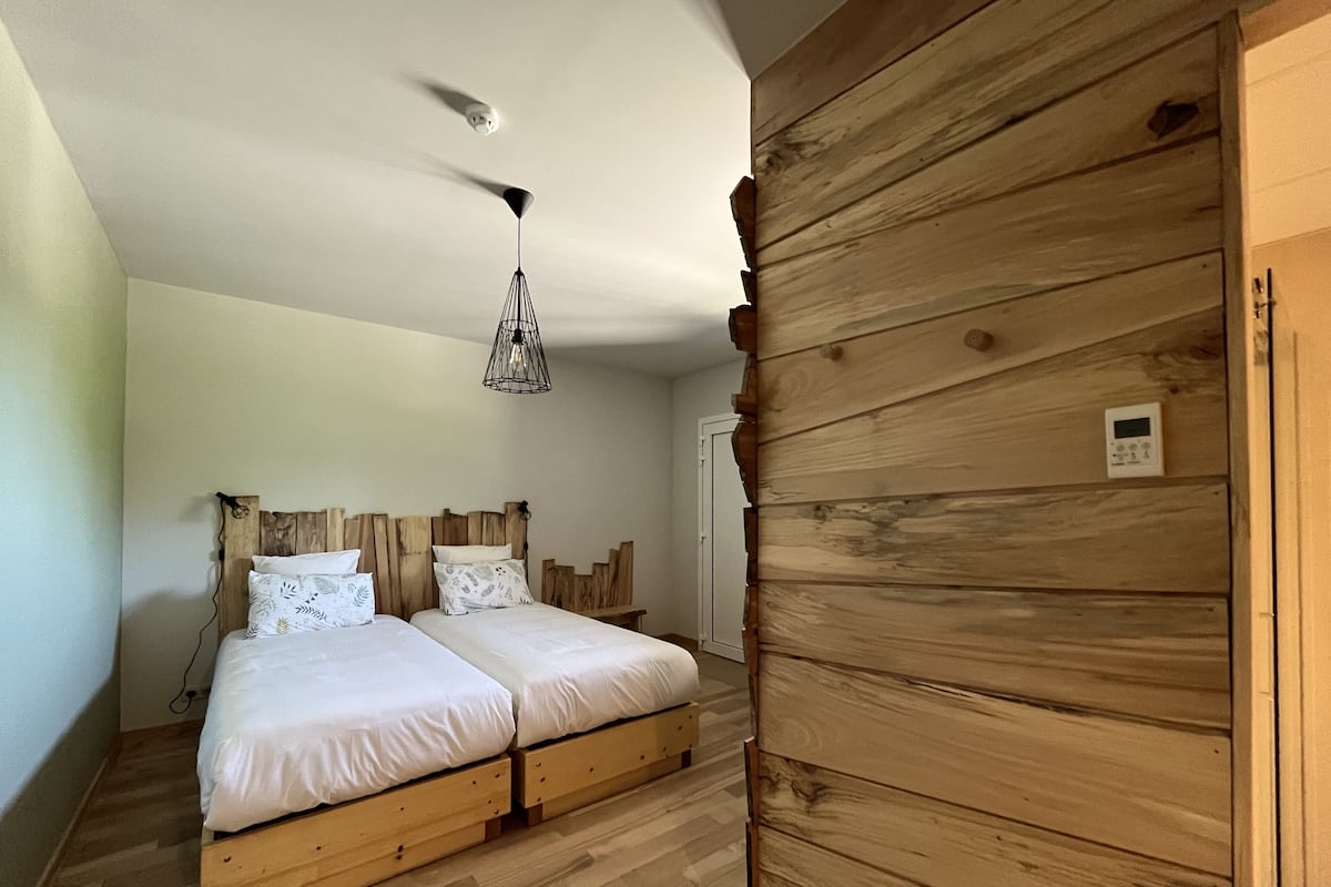Une chambre avec une tête de lit en bois, deux lits, un luminaire suspendu et un thermostat fixé au mur.