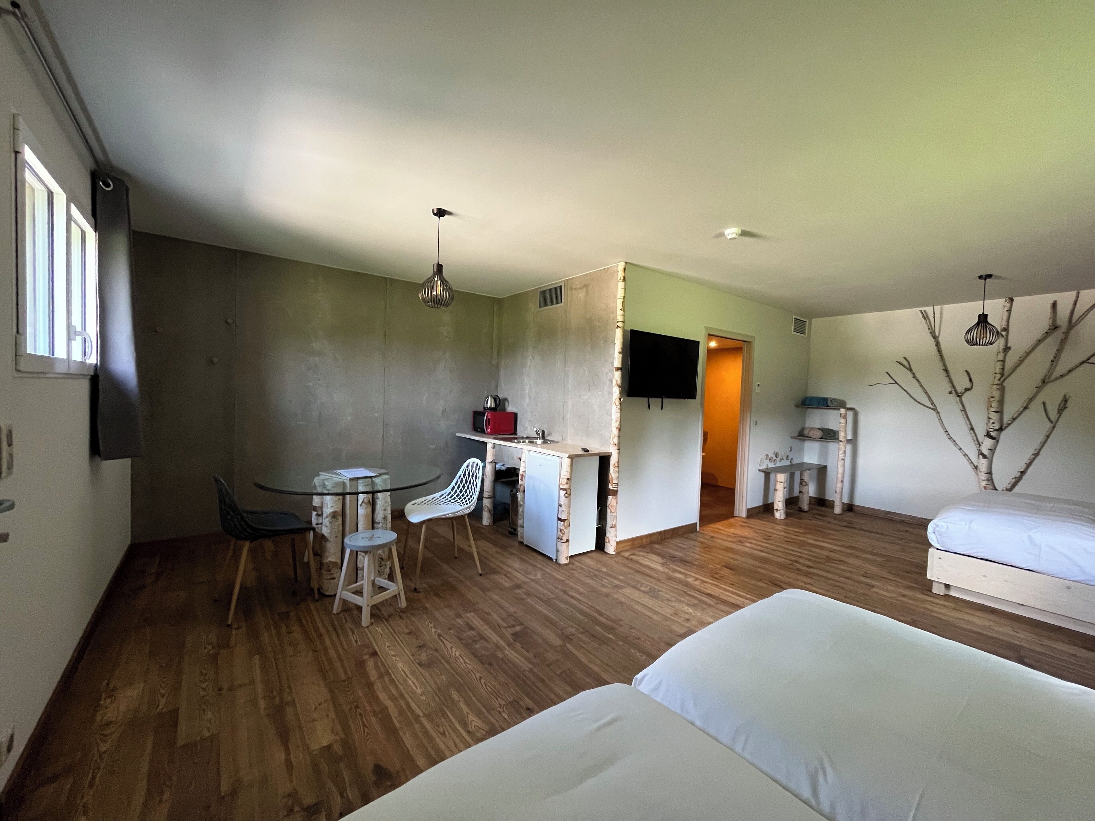 Una habitación compacta con una cama, un pequeño espacio kitchenette, una zona de comedor con mesa y sillas, y un televisor montado en la pared.