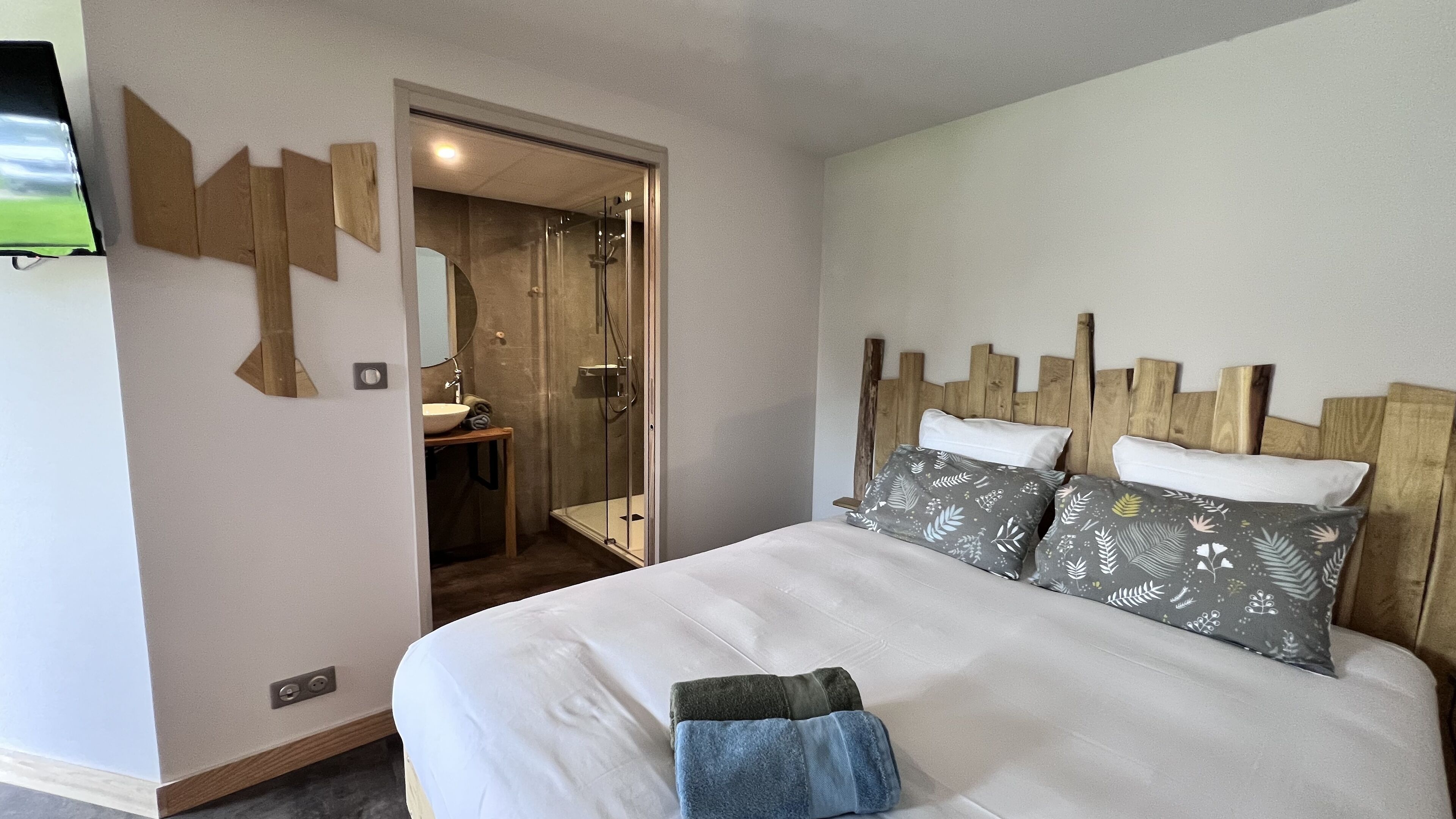 A hotel room with a bed, a wooden headboard, a bedside table with a mirror, and a shower area.