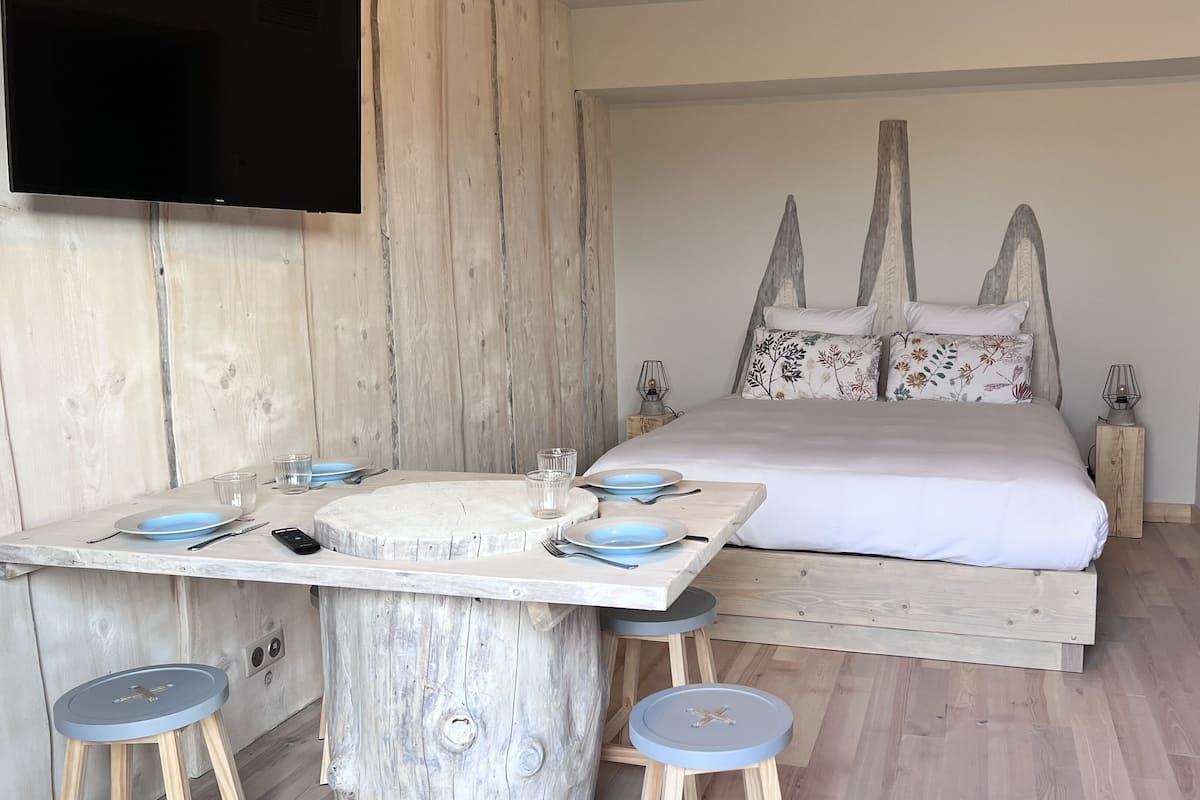Une chambre à coucher avec un lit, une tête de lit en bois, une table de chevet avec une lampe, et un coin repas avec une table dressée pour deux personnes.