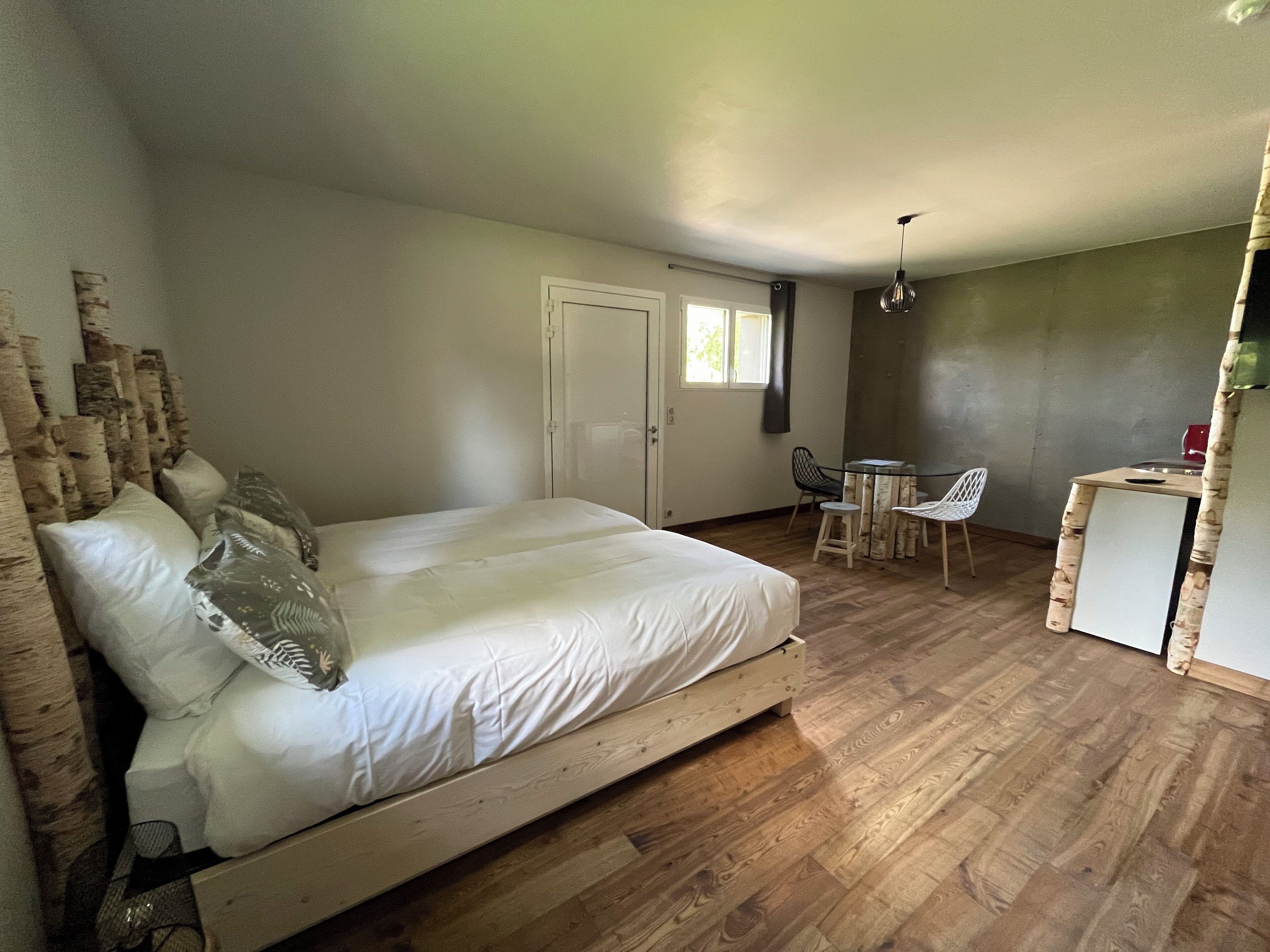 A bedroom with a wooden bed, a small dining area with a table and chairs, and a kitchenette.
