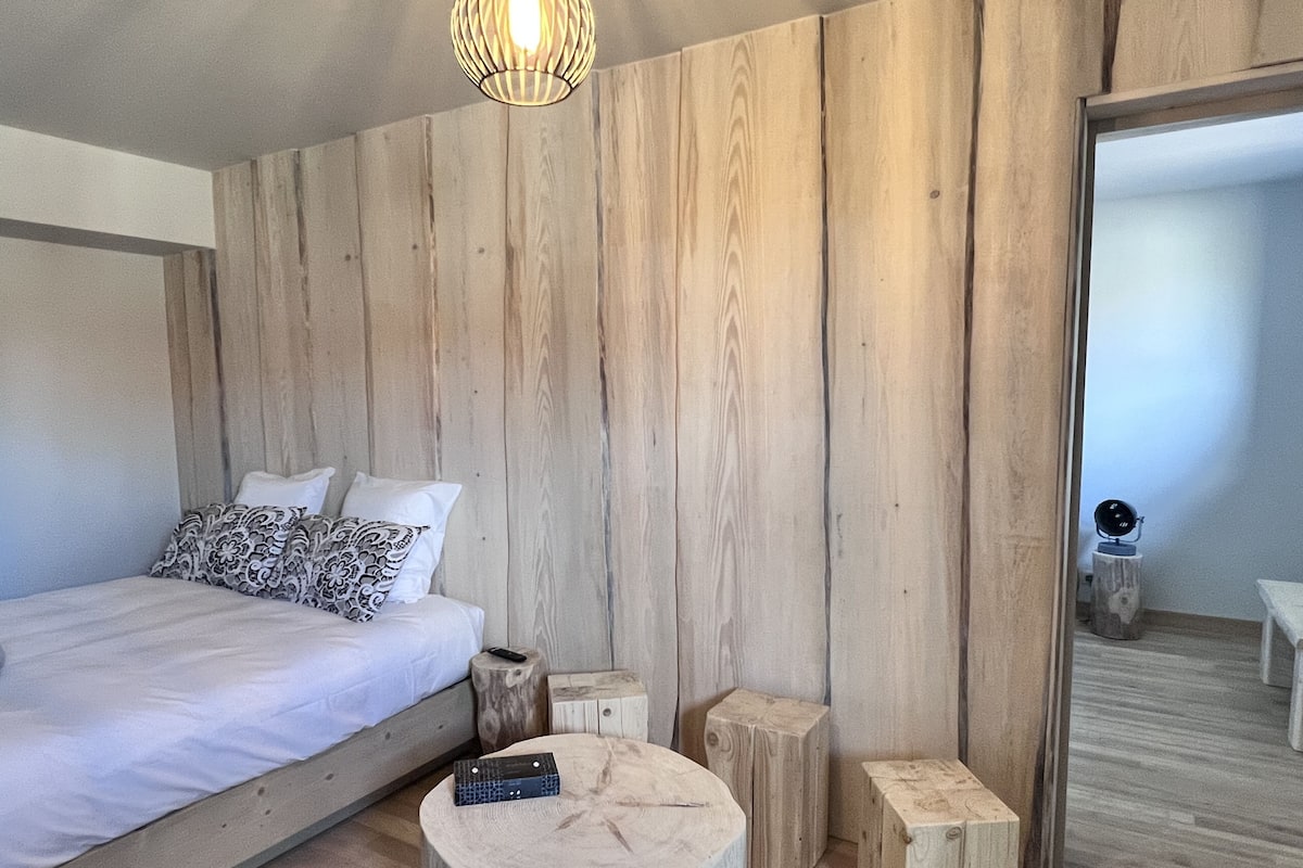 Une chambre avec un mur en bois, un lit agrémenté d’oreillers à motifs, un petit tabouret rond en bois et une petite table avec une télécommande.