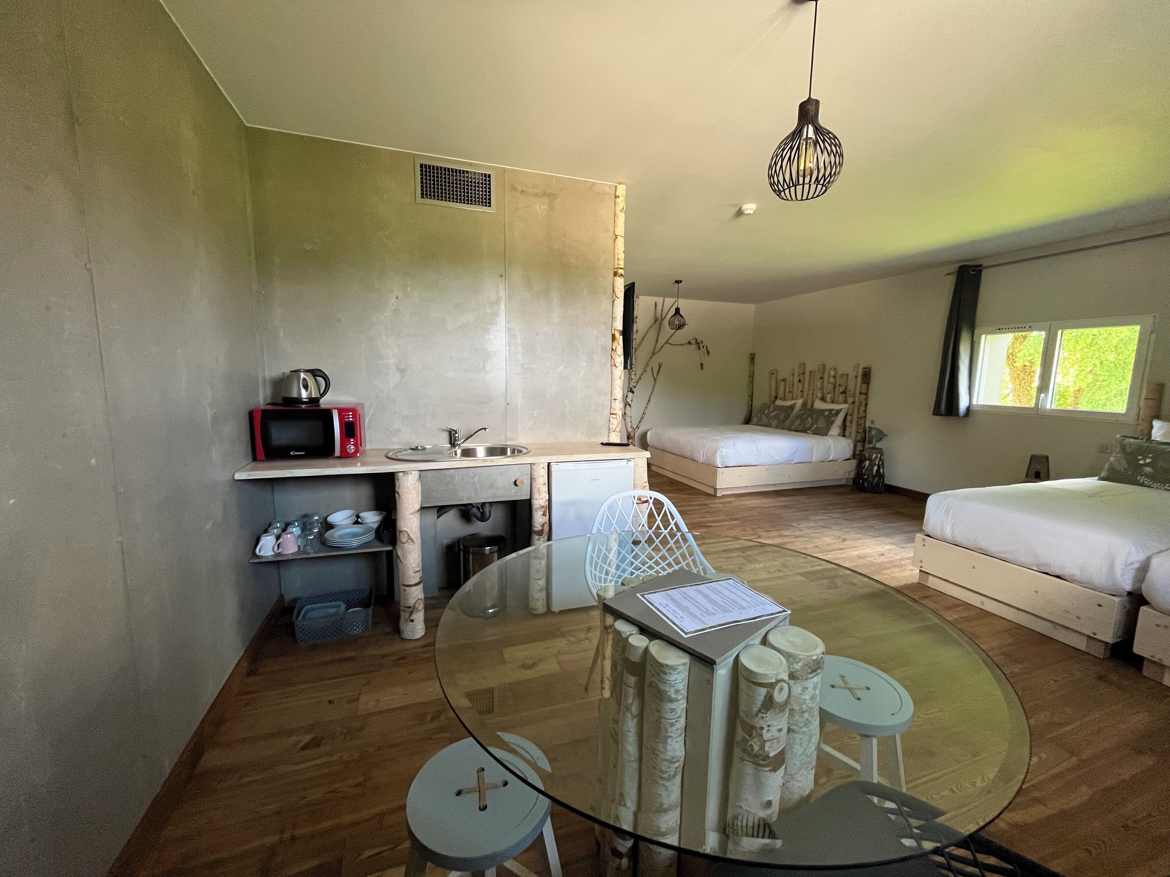A modern bedroom with a glass dining table, a white sofa, a bed, and a kitchen area with a microwave and sink.