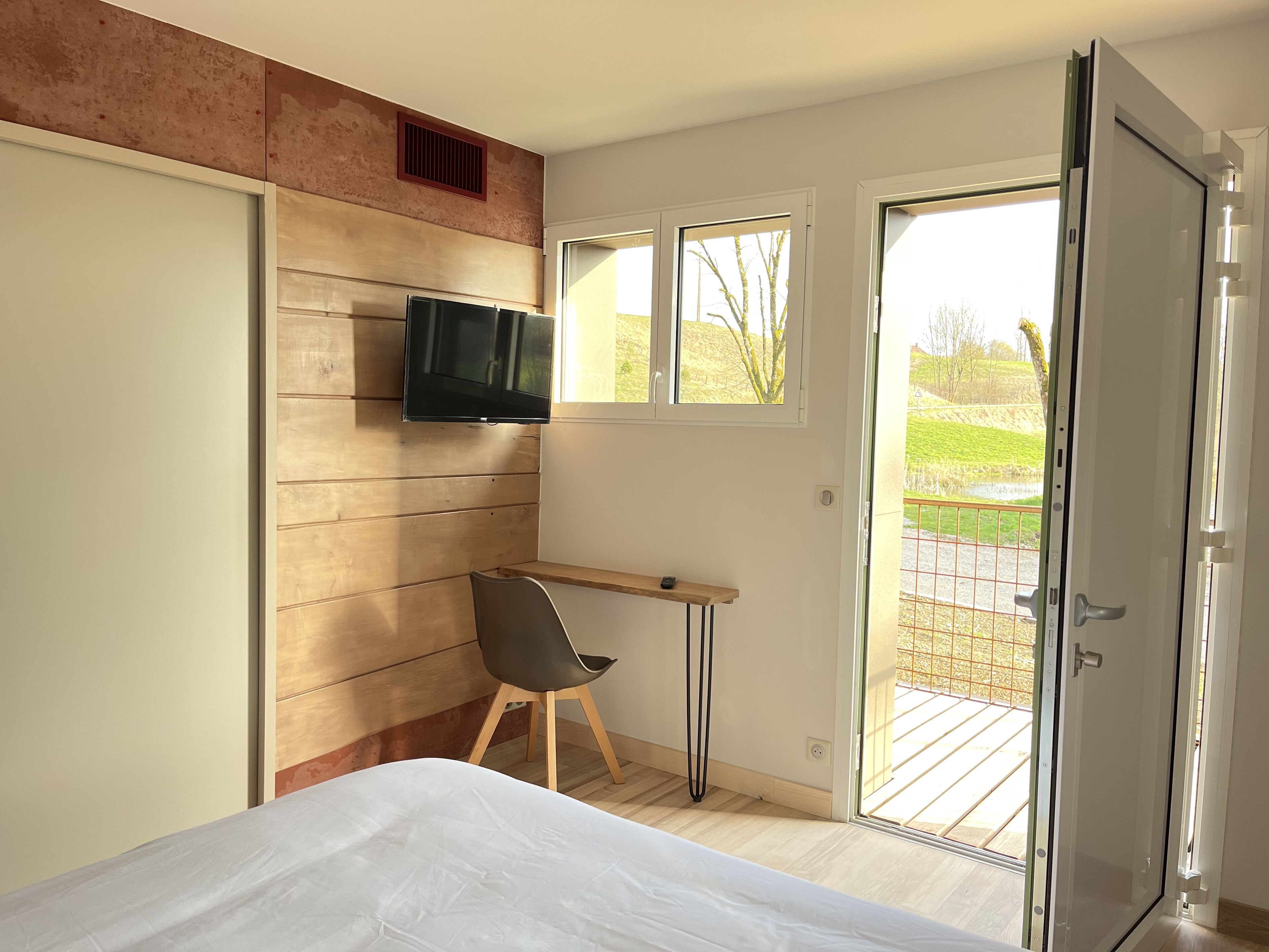 Een slaapkamer met een bed, een bureau, een stoel, een televisie en een schuifpui naar een balkon.