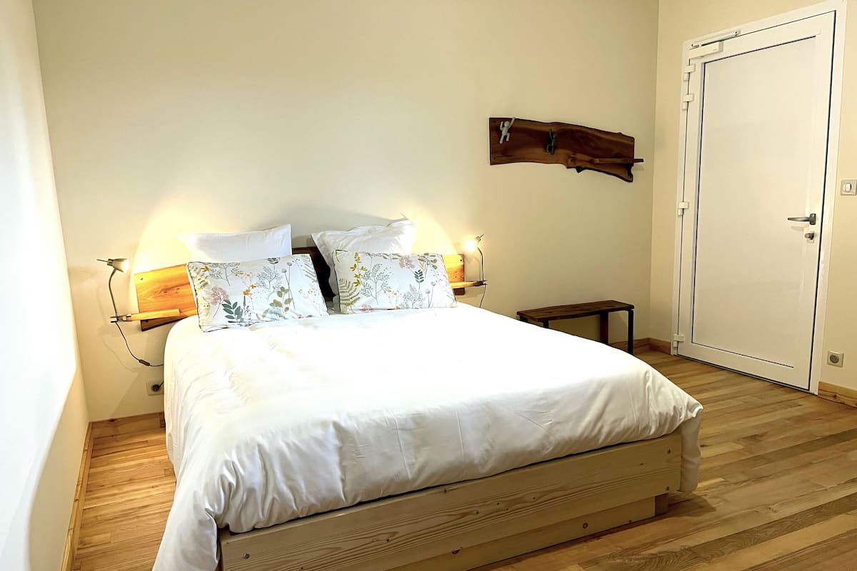 Un lit bien fait, avec du linge de lit blanc et des coussins à motifs floraux, une table de chevet avec une lampe et un banc en bois.
