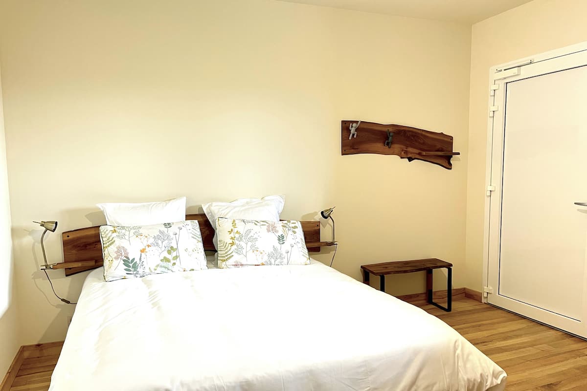 Une chambre à coucher avec un lit en bois, du linge de lit blanc, une petite table de chevet et une tête de lit en bois agrémentée d’un élément décoratif.